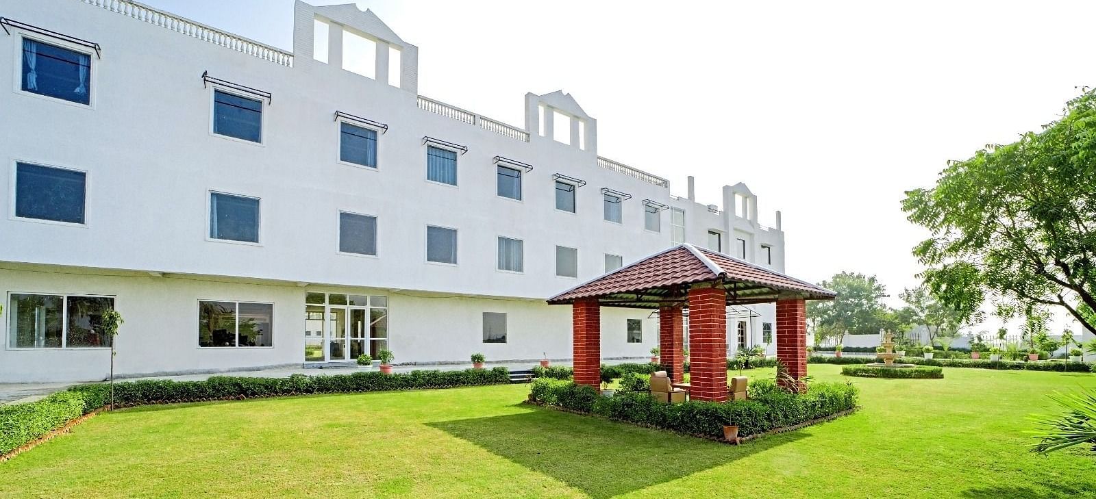 A lawn with a small red gazebo in front of a modern white building at Grassfield Riveria Resort, Jaipur.