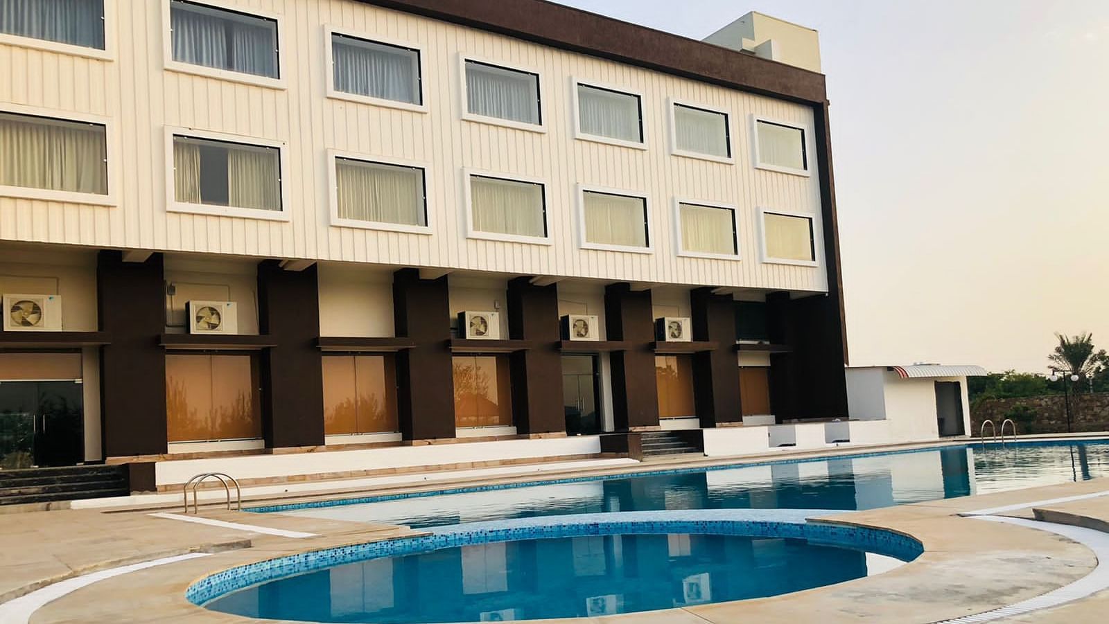 The exterior of the Bamboo Saa Mulberry Resort, Pushkar, featuring a multi-story building with a light-coloured facade with a swimming pool is visible in the foreground.
