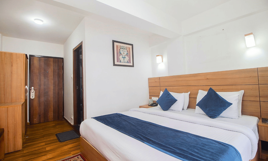 The Deluxe Room with double bed, framed art work on the wall, TV, Wardrobe, Dustbin, Table and desk with two water bottles on the top at  Central Heritage Resort and Spa (Formerly Fortune ITC Resort) The Mall Ro