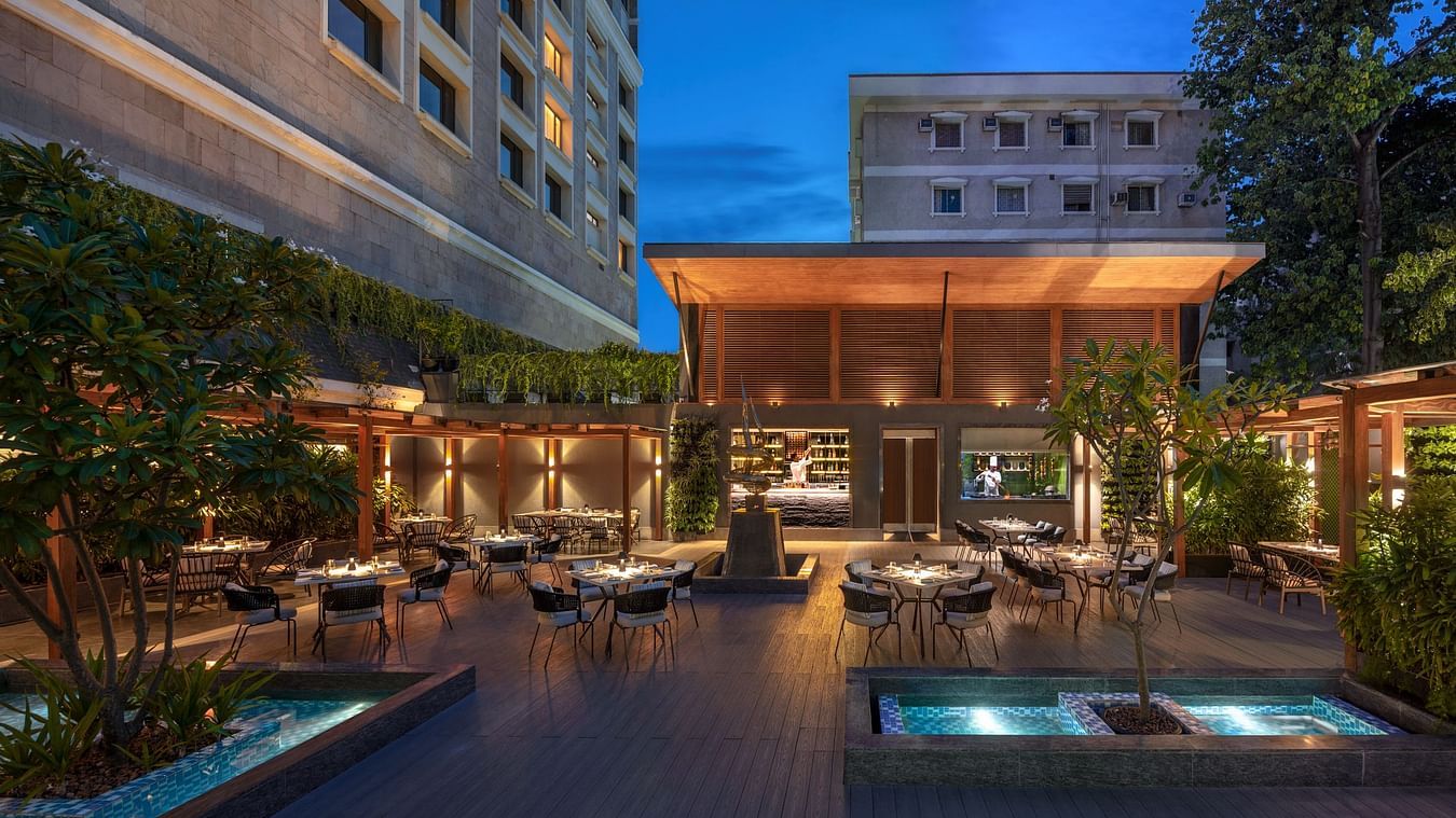 An outdoor dining area with tables, chairs, and plants, illuminated by warm lighting along with swimming pool. 