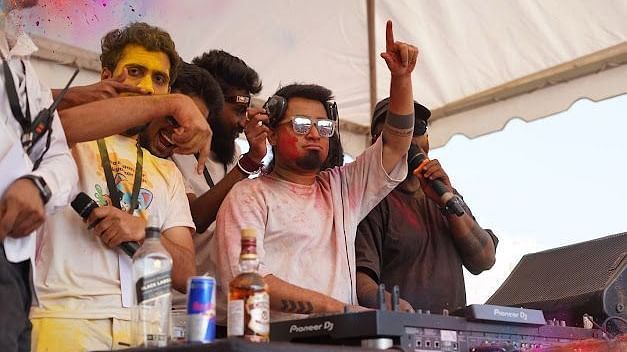 DJ performing on stage with energetic crowd enjoying Holi festival, music, and vibrant colours.