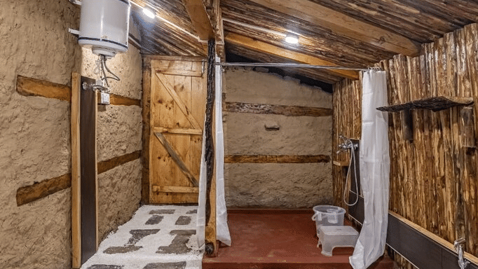 The bathroom of the Deo Tibba Cottage featuring a bath area separated with white curtains along with a water geyser at Amara Upepo - The Sky Village, Manali.