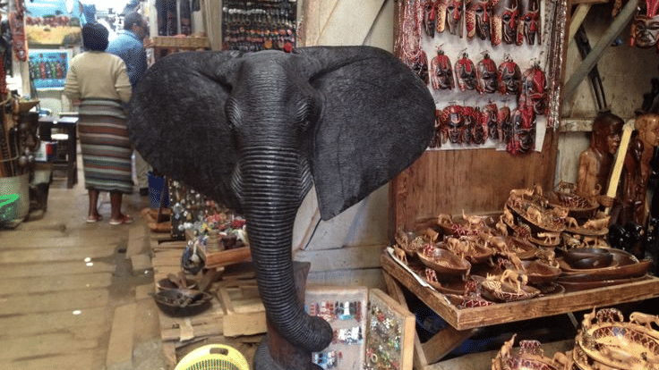 A large dark sculpture of an elephant head stands in the middle of an outdoor market stall surrounded by many small wooden carvings and woven items.