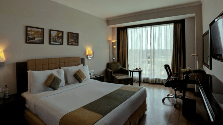 Modern hotel bedroom at Goldfinch Bengaluru with a large bed, brown runner, work desk, chair, wall art, and a large window.