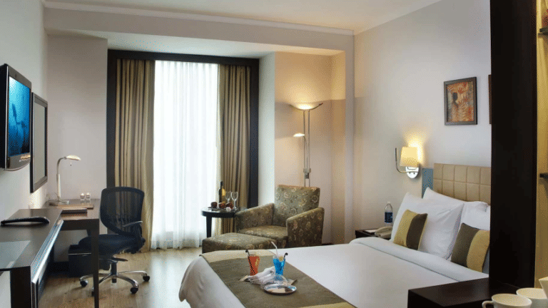 Bed with a runner, side tables, armchair, and a work desk in a brightly lit guest room at Goldfinch Bengaluru.