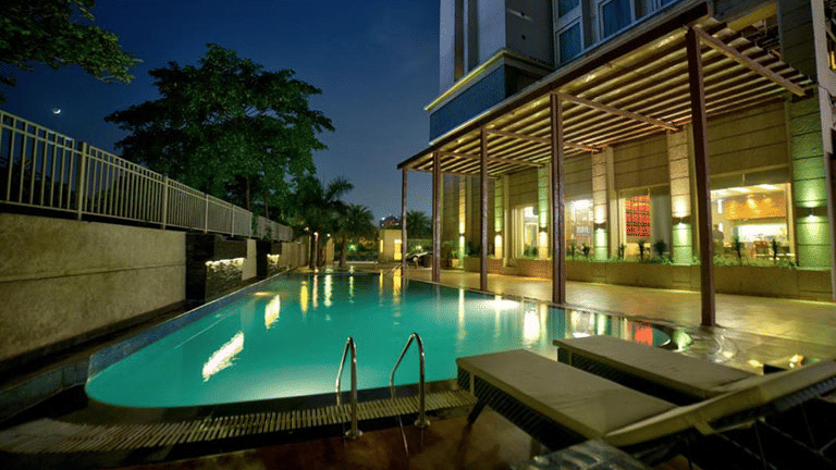 Outdoor swimming pool at night with poolside lounger chairs and wooden trellis architecture at Goldfinch Bengaluru.