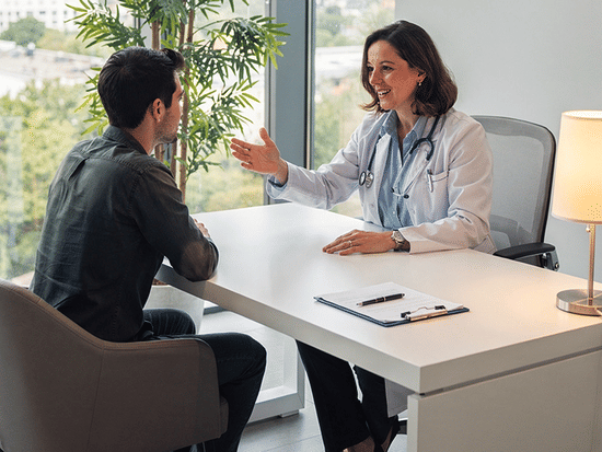 A  healthcare professional in white coat speaking with patient across desk, clipboard and lamp, bright window view with greenery.