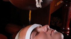 A woman lies with eyes closed as warm oil streams from a hanging pot onto her forehead during a Shirodhara Ayurvedic treatment at Ayur Ashram