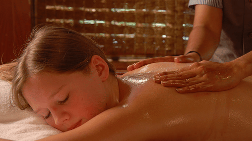 A woman lying on her stomach with her eyes closed receiving massage with oil on her upper back at Uday Suites