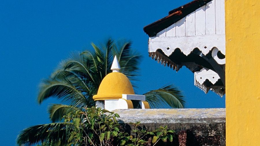 A bright blue sky above the yellow and white architecture of Fort Tiracol, Goa, with palm trees framing the view.