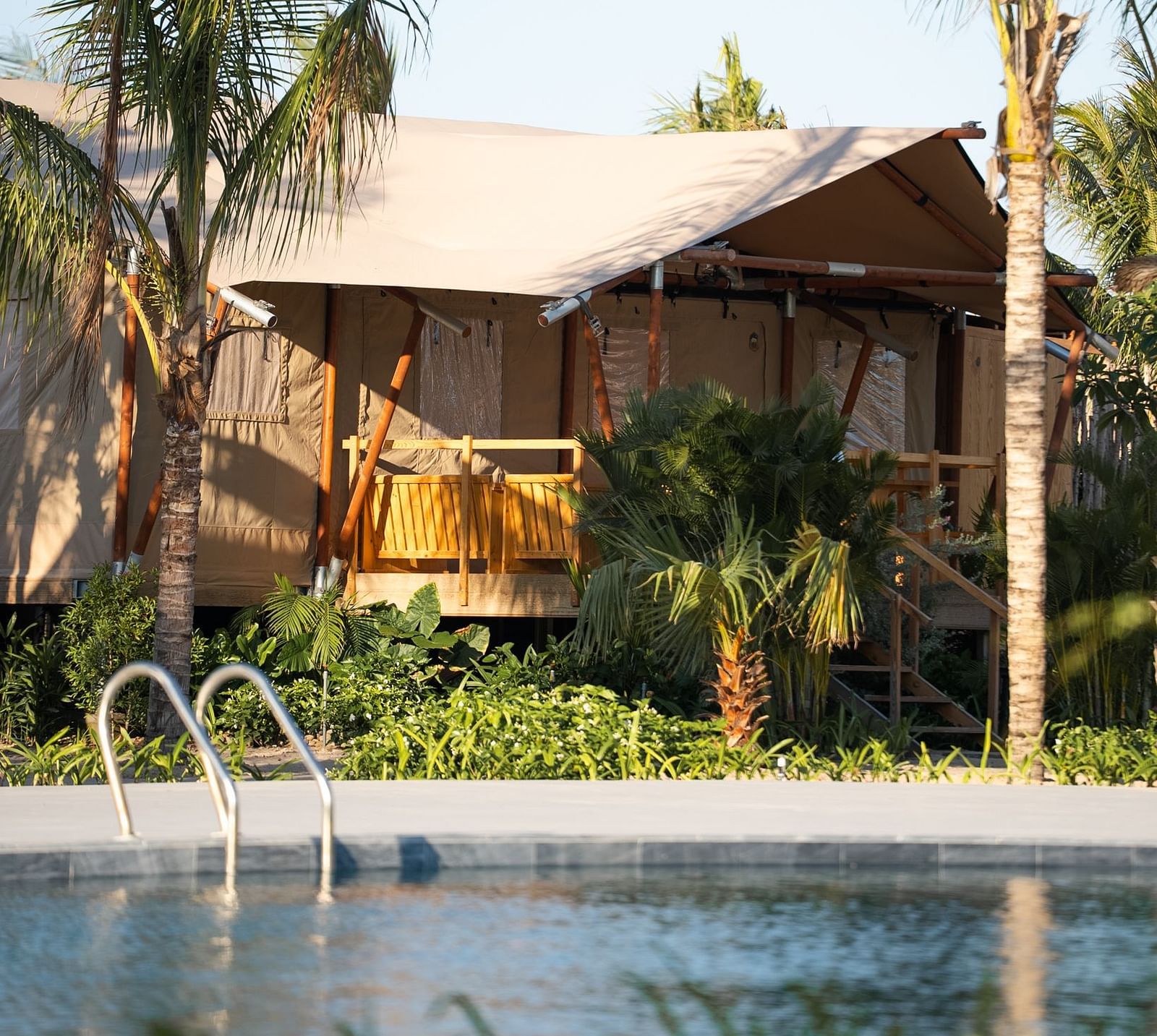 The Grateful Tent at WoCAL Retreat Ke Ga overlooking a pool, with a wooden porch and steps leading into a spacious outdoor seating area, surrounded by towering palms and open sky.