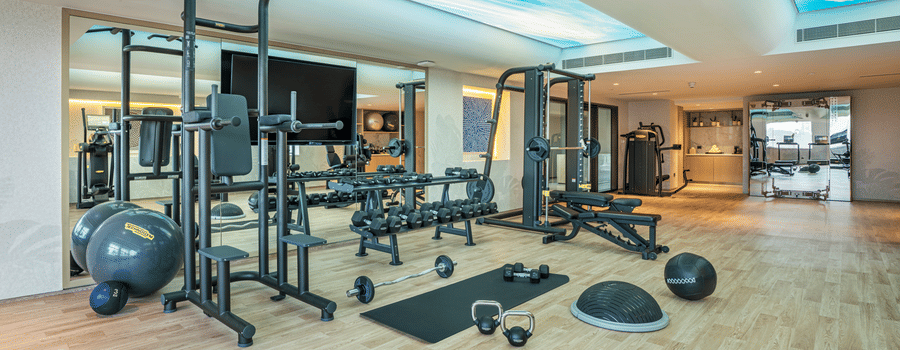 A gym at The Residency Towers Chennai featuring weight racks and exercise equipment with mirrored walls, and ceiling with the sky visible through the glass