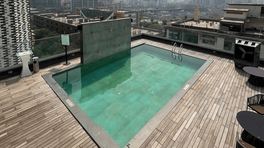 Angled view of a square rooftop swimming pool with green water on a wooden deck overlooking the city.
