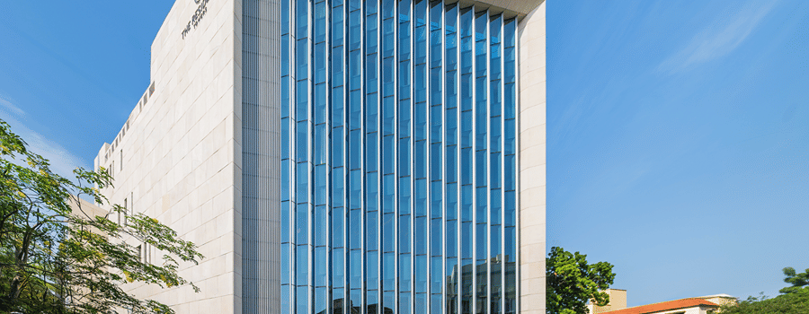 Hotel facade in daylight with white stone cladding, vertical blue glass fins, a stone boundary wall and trees by the driveway at The Residency Towers Chennai.