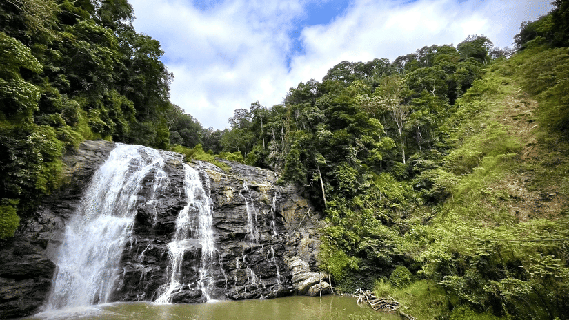 A waterfall flowing over a rocky cliff surrounded by trees and green vegetation under a partly cloudy sky.