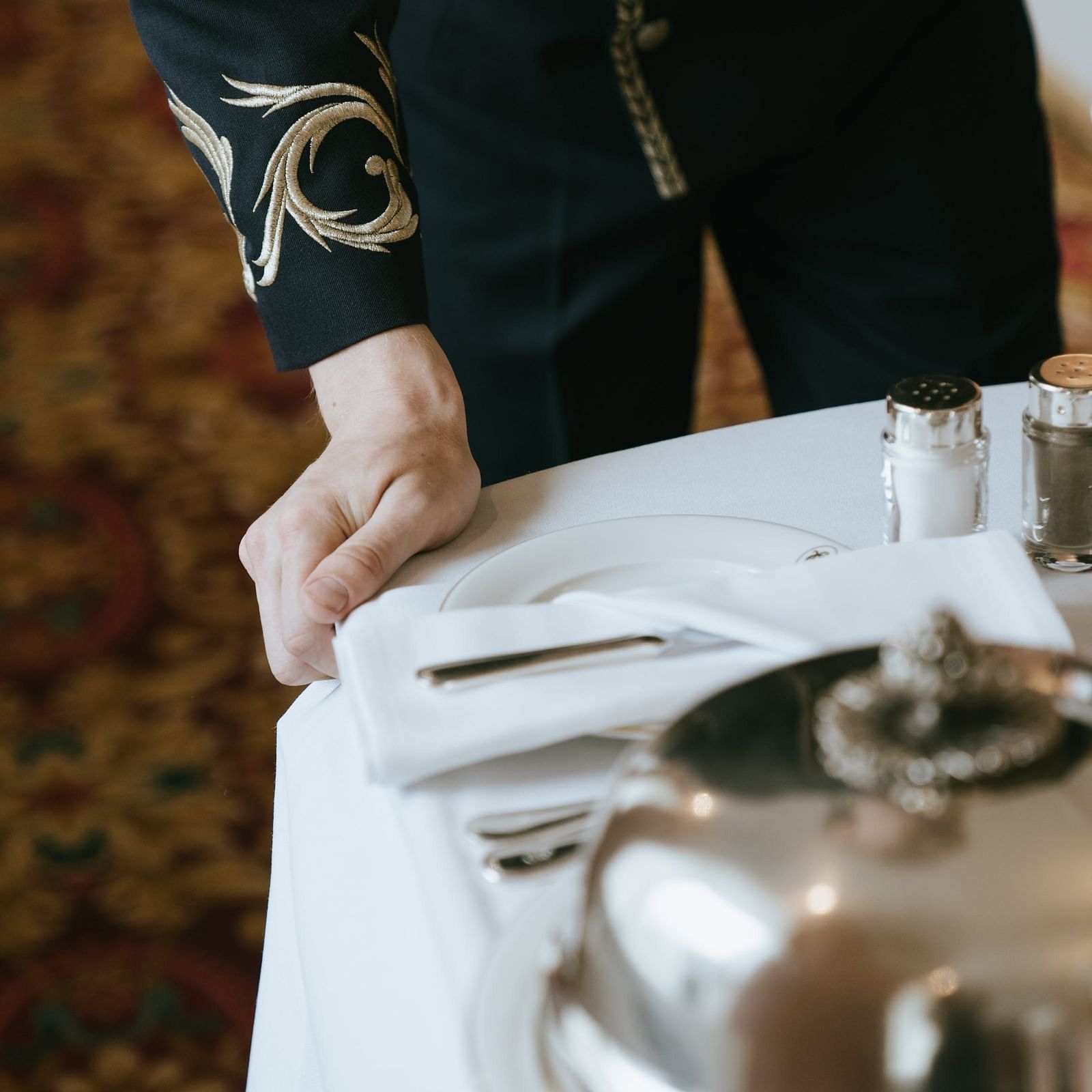 A waiter in a formal uniform preparing a dining table with a silver cloche, salt and pepper shakers and cutlery on a white linen