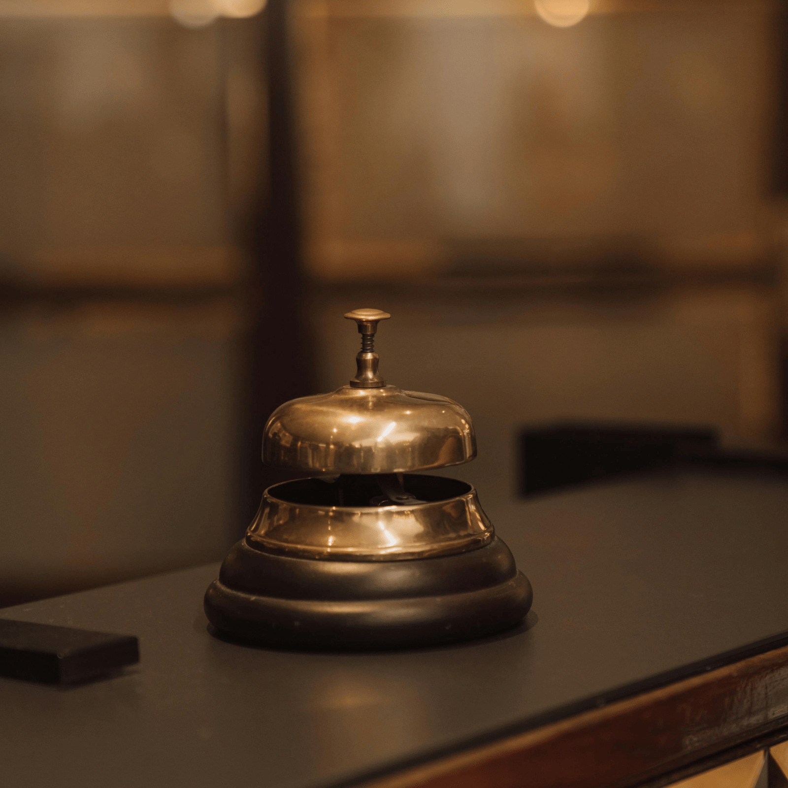 A close-up of a vintage brass service bell with a wooden base, sitting on a dark wooden counter, likely at a hotel reception.
