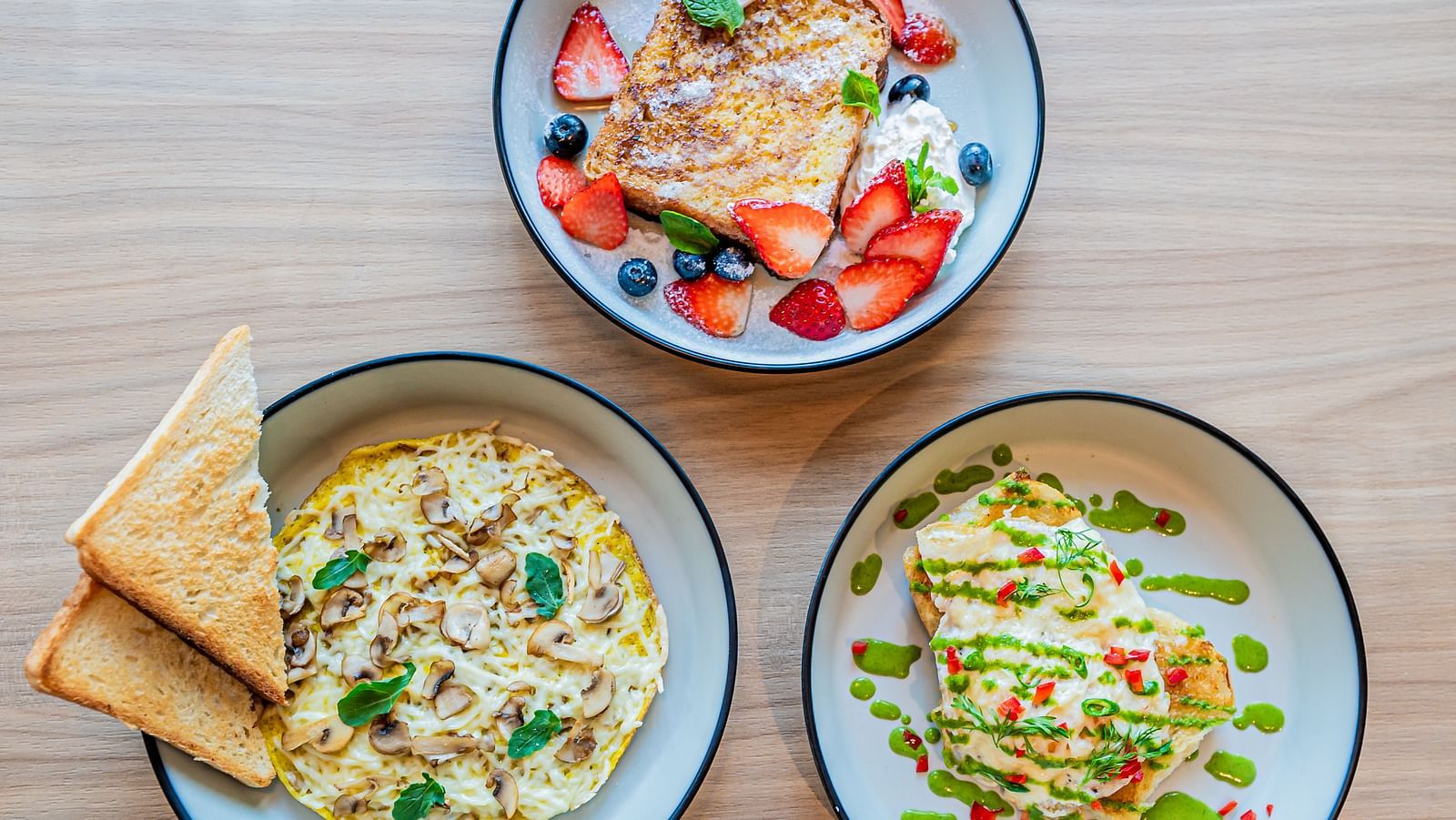 Three plates at Indie Stays Mumbai International Airport: one with mushroom omelette and basil leaves, one with toast topped with strawberries and blueberries, and one with egg toast and veggies.