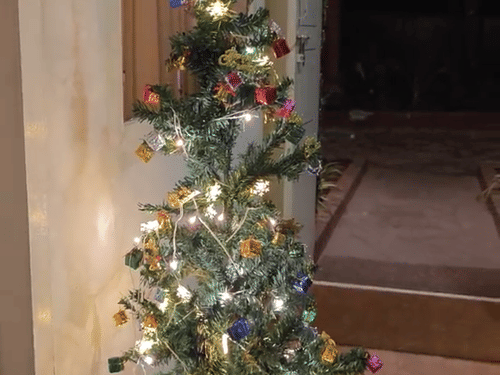 A vertical shot of a small, illuminated Christmas tree with colorful ornaments standing in a pot on a light-colored floor, against a plain wall.