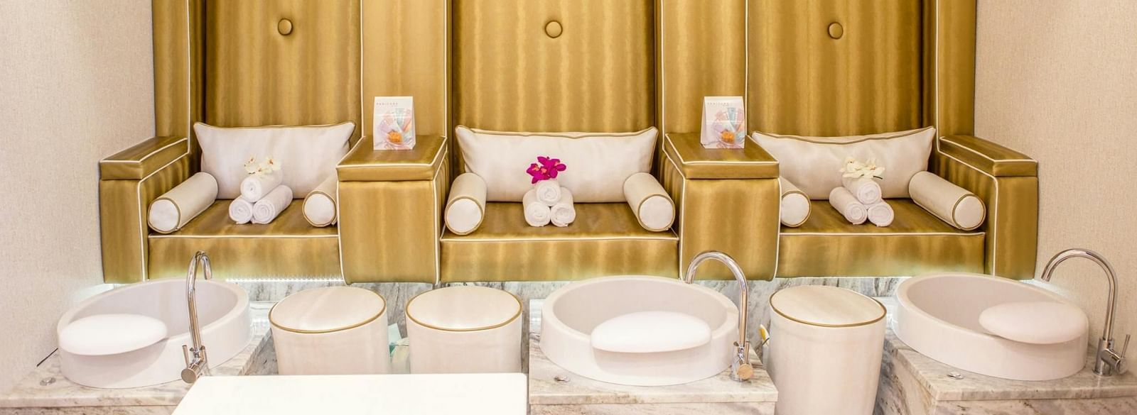 A row of elegant chairs with cushions and individual foot basins in a modern, brightly lit spa treatment room