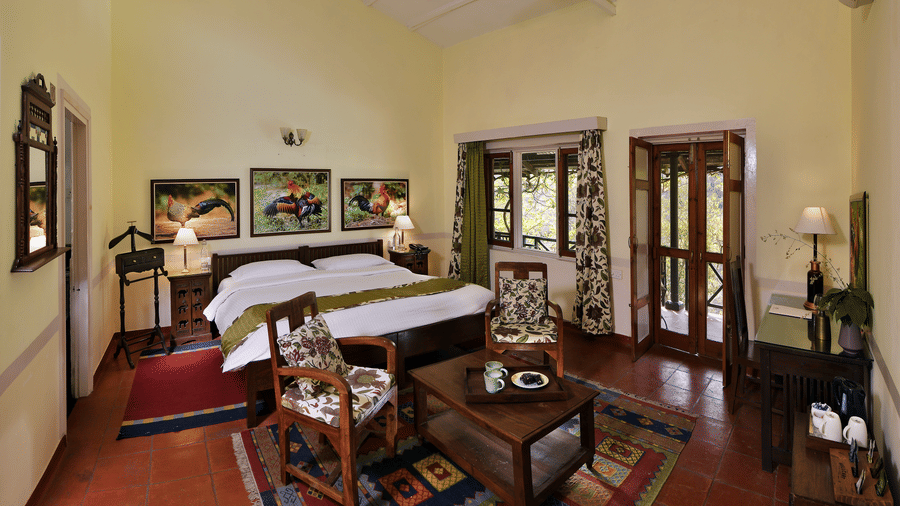 Spacious heritage style suite with a large bed wooden furniture seating area patterned rugs tall ceiling fans and natural light creating a calm and comfortable stay experience at Neemrana’s Glasshouse on The Ganges surrounded by a peaceful indoor atmospher