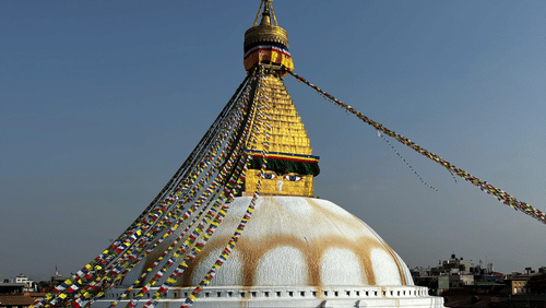 The Boudhanath Stupa featuring a white dome, a golden spire with painted eyes, and colourful prayer flags against a cloudless sky.