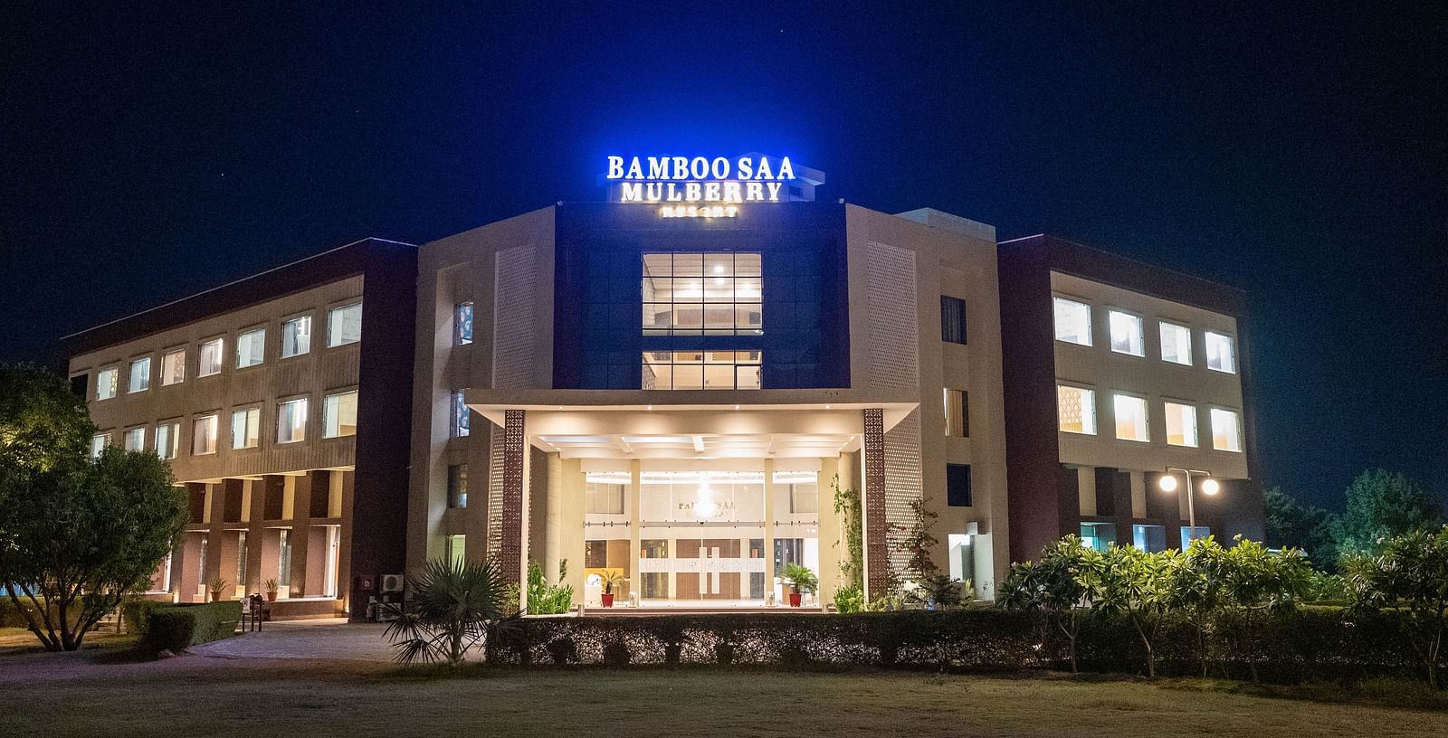 Front view of the grand facade and portico of Bamboo Saa Mulberry Resort, Pushkar with a large illuminated logo at night.