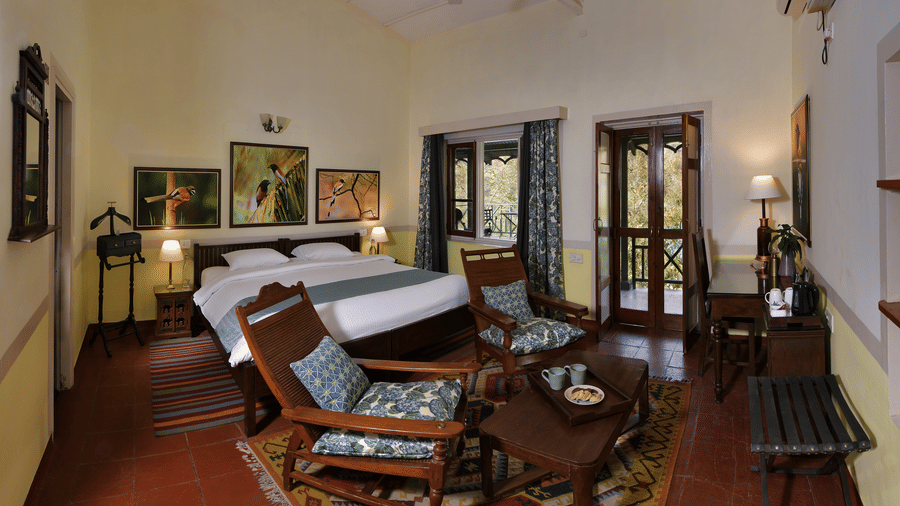 Elegant premium suite bedroom with wooden bed side tables seating corner ceiling fans and warm lighting providing a refined and restful stay at Neemrana’s Glasshouse on The Ganges with generous space for extended comfort