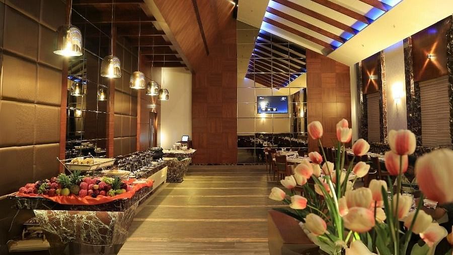 A view of the Aroma restaurant in Dhanalakshmi Srinivasan Hotels, Perambalur, featuring a buffet counter on the left and dining tables in the background. The restaurant has wooden ceiling beams and a floral arrangement in the foreground.