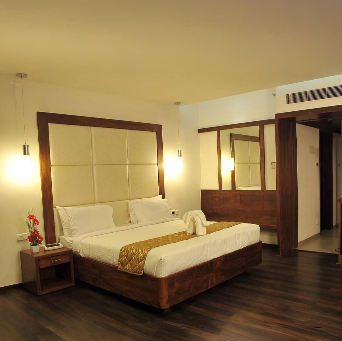 A room with a queen size bed, two side tables, wooden flooring, two mirrors, hanging lights, and a counter with bottles and appliances at Dhanalakshmi Srinivasan Hotels, Perambalur.