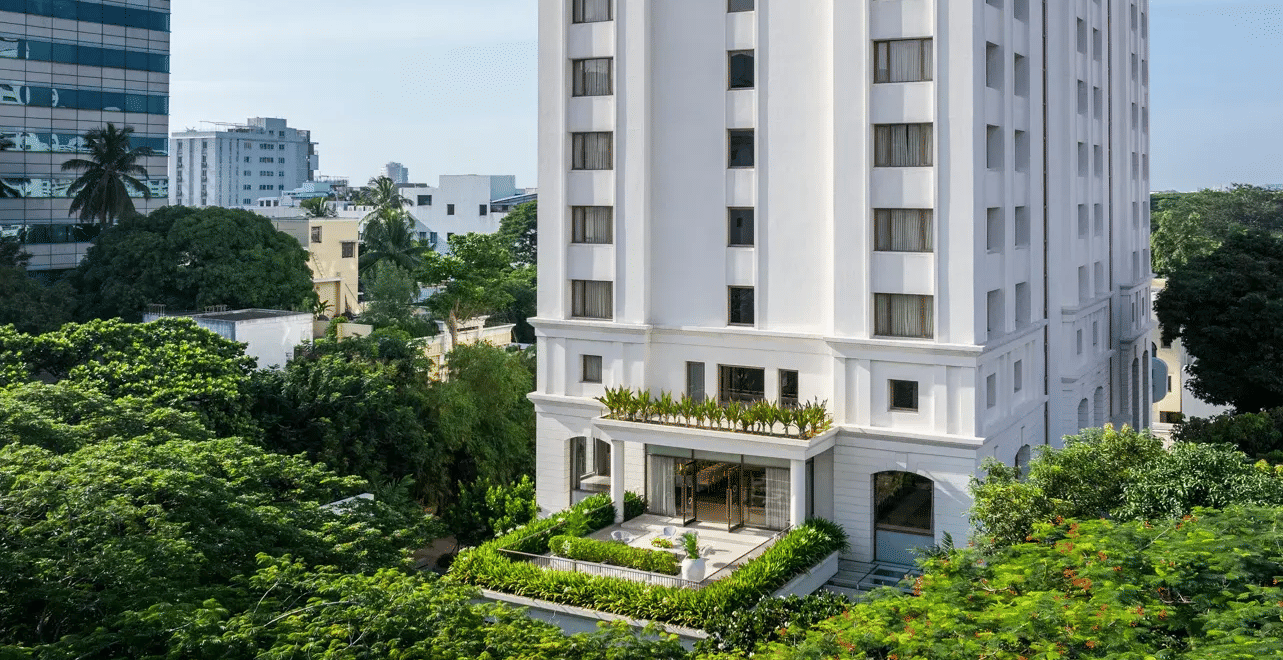 Tall white multi-storey building with arched top-floor windows and a rooftop pergola terrace, seen from below amid trees and the city skyline.