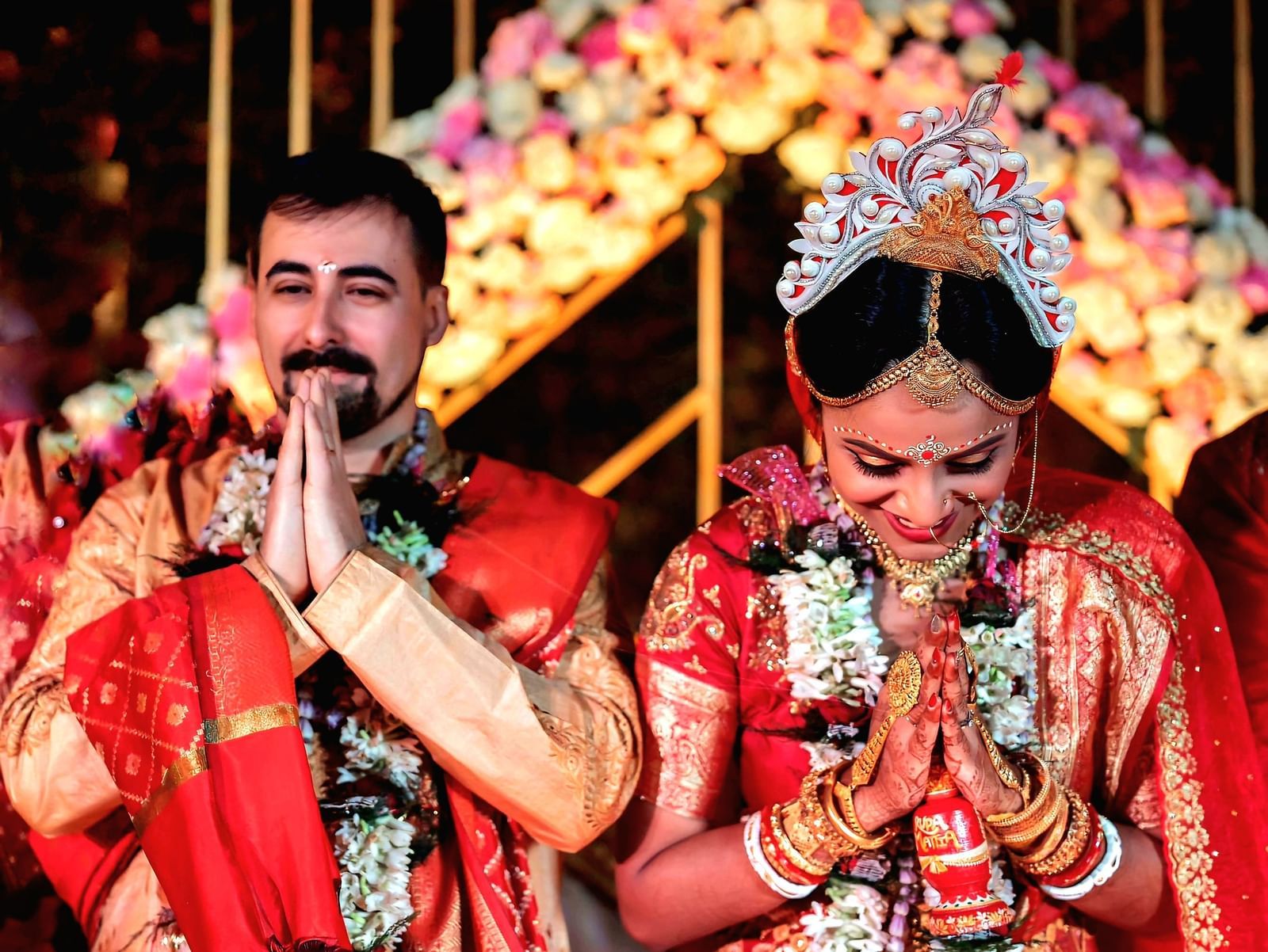 A couple in traditional Indian wedding attire performs a ritual with hands joined under a floral archway