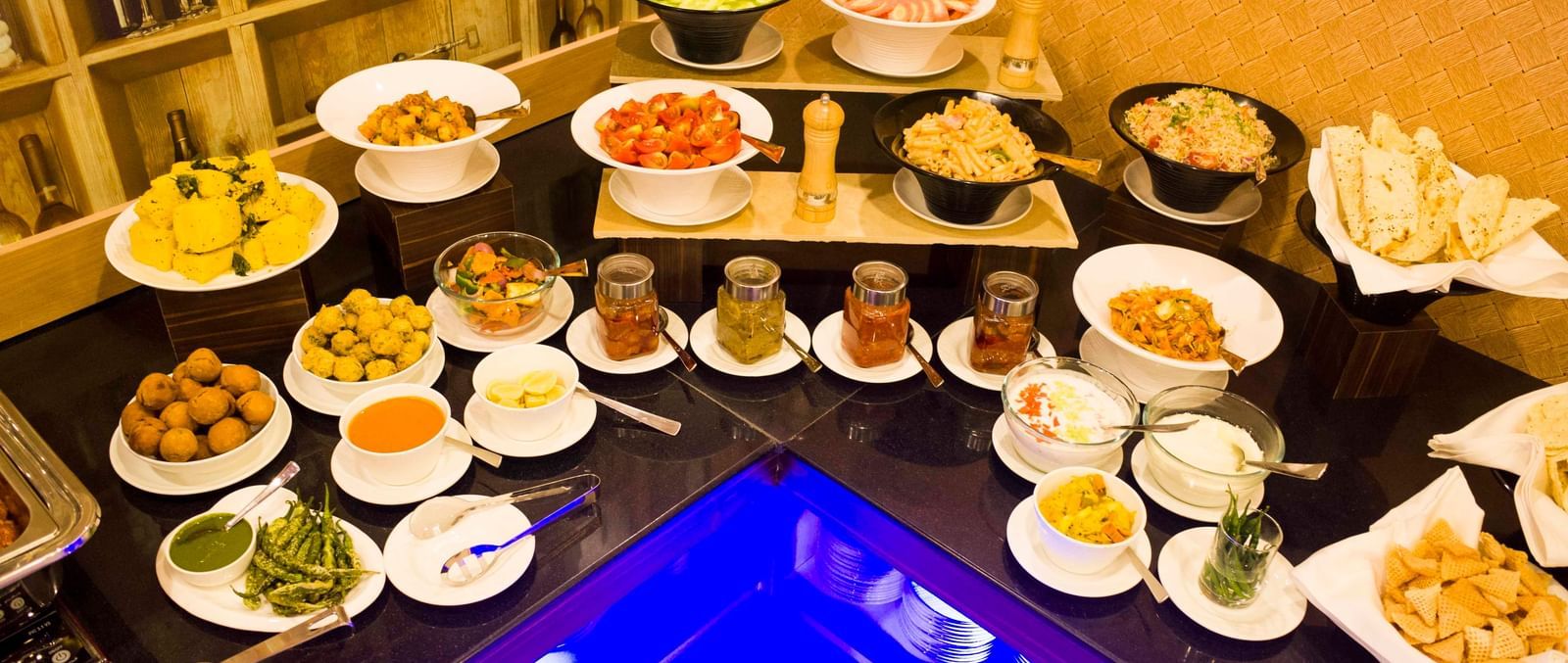 A lavish buffet spread with a variety of appetizers, salads, and condiments, featuring a blue-lit counter element | Daksh The Madhuvan Suites