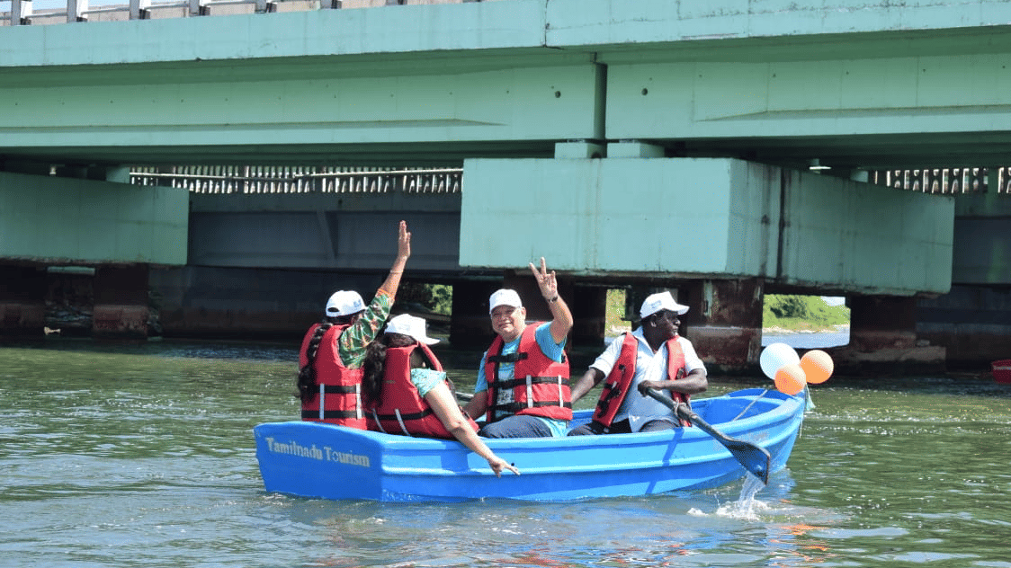 People wear life jackets and ride in a boat on the water. A bridge structure spans across the background above them.