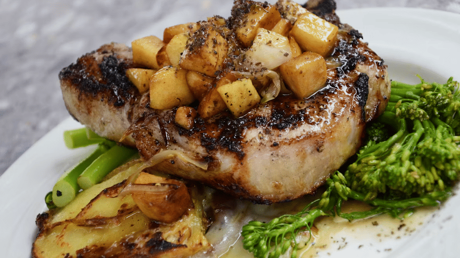 Grilled meat chop topped with sautéed apple cubes, served with broccolini, green beans, and roasted potatoes on a white oval plate.