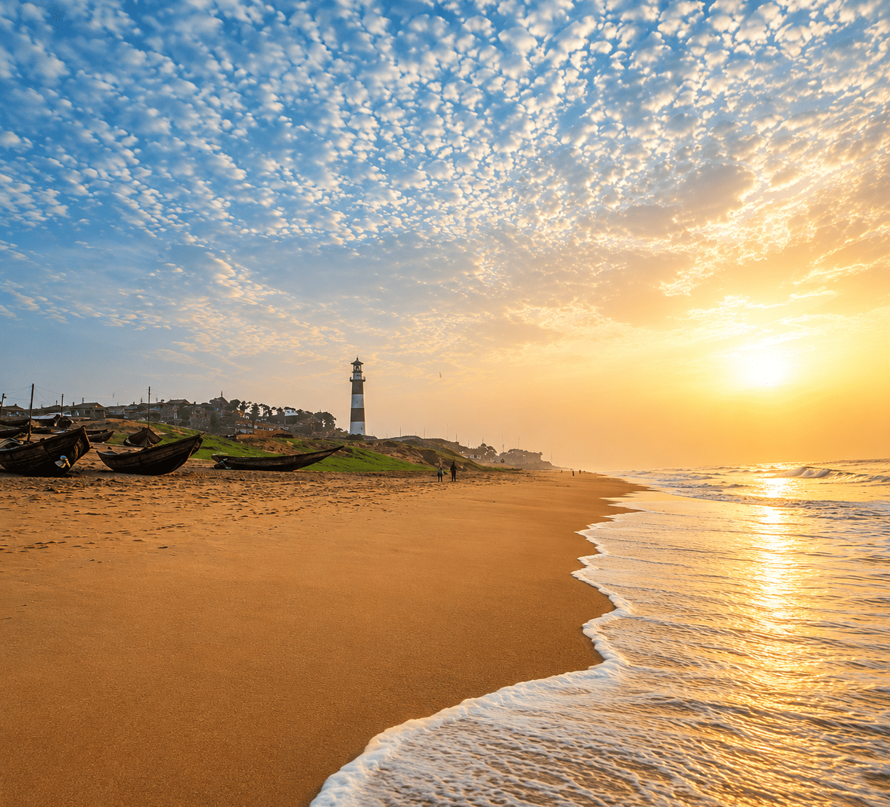 Sunrise over Gopalpur Beach with wooden fishing boats on the sand, a striped lighthouse and shoreline houses, near MAYFAIR Palm Beach Resort, Gopalpur-on-Sea.