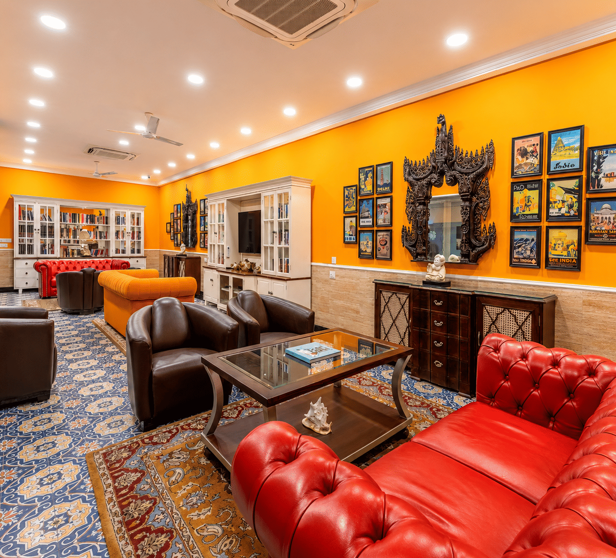 A lounge area at MAYFAIR Palm Beach Resort, Gopalpur-on-Sea. Sofas and armchairs surround a central coffee table on a patterned carpet. Framed pictures hang on the wall, and a cabinet sits in the background with lot of books in the shelves.
