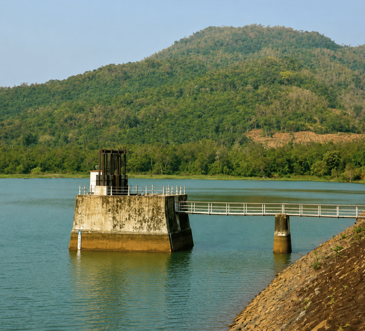 Salia Dam reservoir with a concrete intake tower, walkway bridge on piers and a forested hill beyond, near MAYFAIR Palm Beach Resort, Gopalpur-on-Sea.