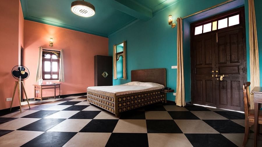A guest room at Tijara Fort-Palace - 19th Century, Alwar features a bed, a mirror, a standing fan, a door with curtains, and checkered flooring.