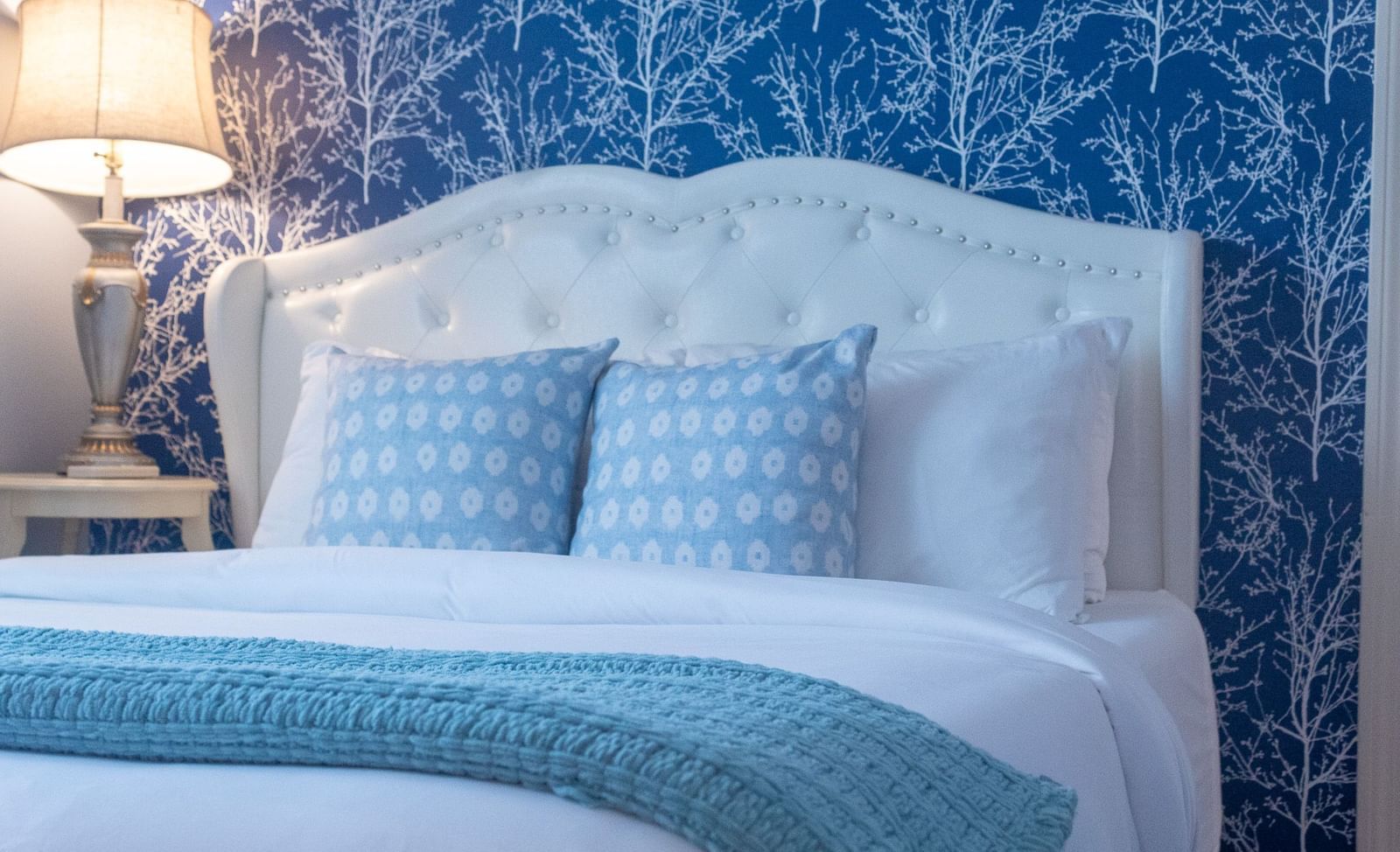 A bedroom with white tufted headboard, navy tree wallpaper, blue pillows and teal throw blanket at Hartstone Inn.