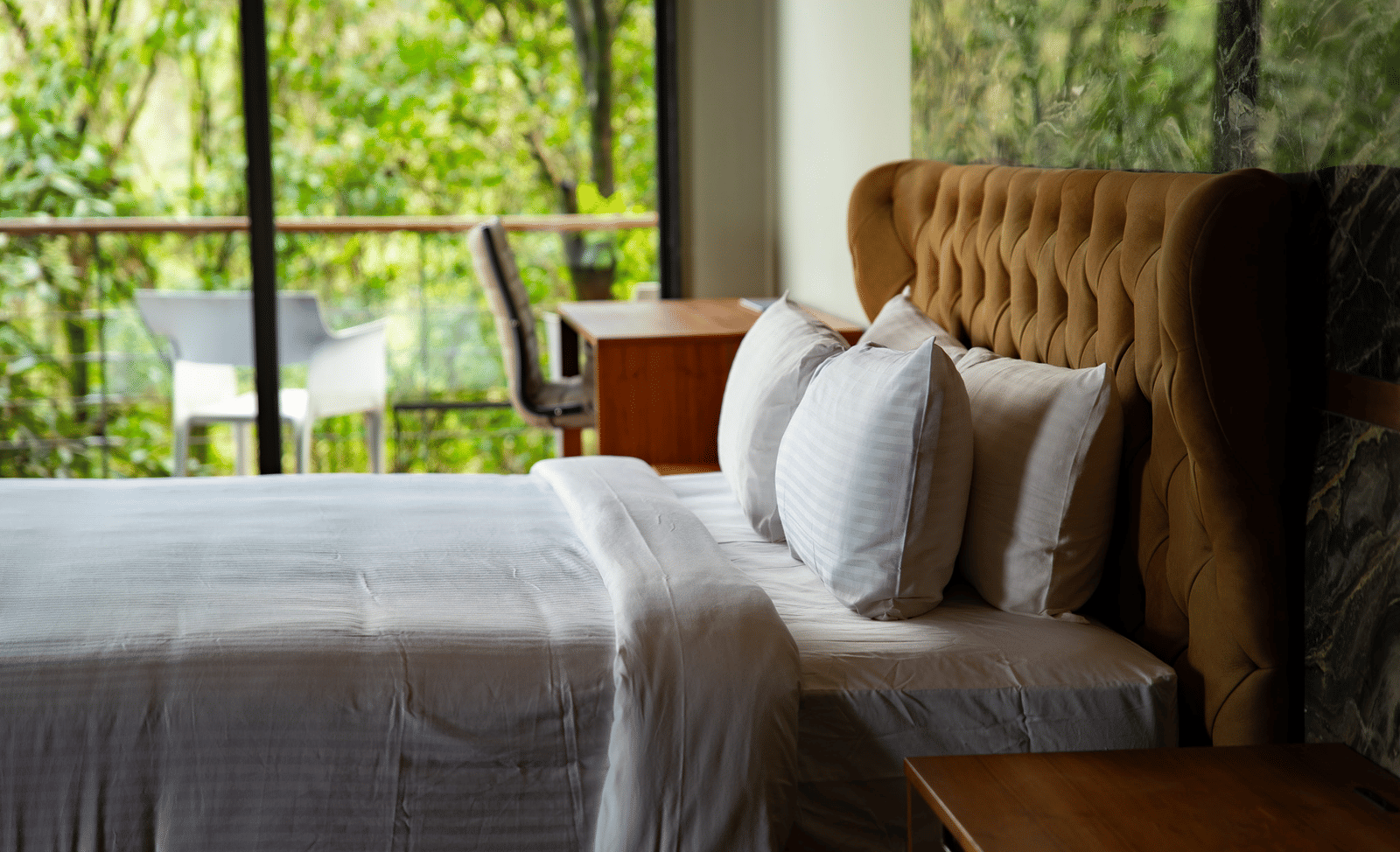 A close-up of a large bed with a high, tufted headboard at Stanley Revelation, with a forest visible outside the window.