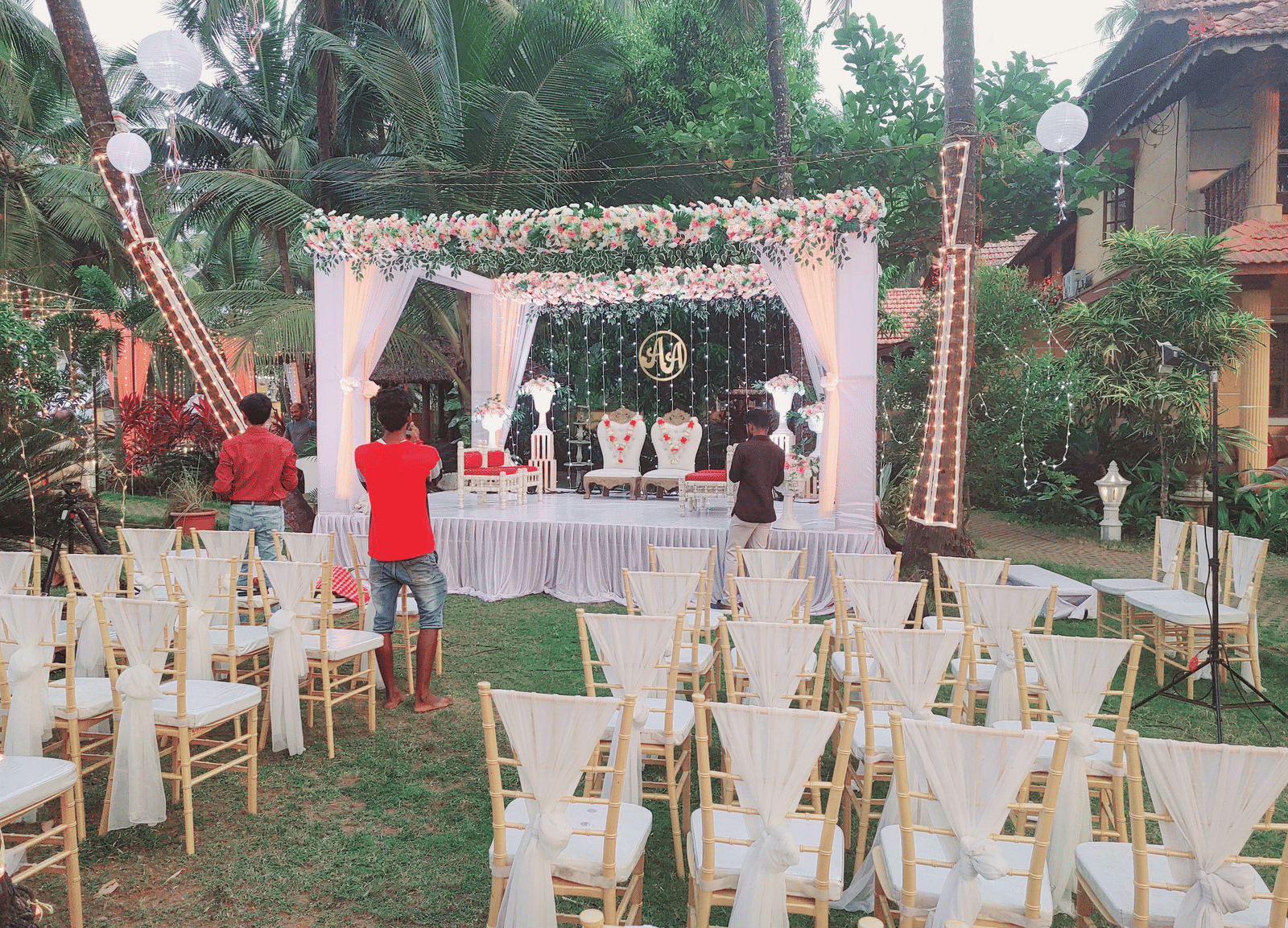 Outdoor wedding setup with white chairs, floral arch, and people arranging decorations at Paradise Lagoon Resort, Udupi.