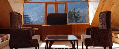 This image shows a cosy seating area in a hotel room with large windows offering scenic mountain views. It features two armchairs, a wooden coffee table, and a warm, wood-panelled interior | DLS La Serene Manali