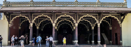 Front view of the Tippu Sultan's Palace in Bengaluru, during a sunny day with a pathway lined with green and yellow-hued bushes.