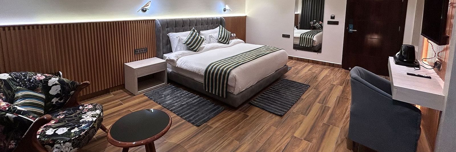 A shot of the deluxe room featuring a bed, bed side tables, two chairs, coffee table, work desk, a couch, a flat screen TV, and a full length mirror - DLS The Emerald, Nainital