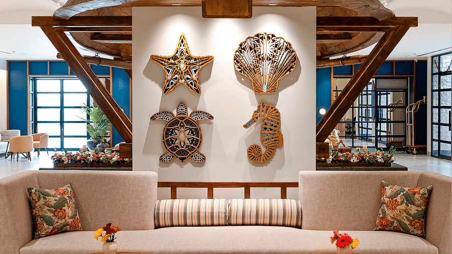 A lounge area features a spacious sofa and a three-legged wooden table with a vase of flowers in the centre. The wall is adorned with sea-themed, wall-mounted decorations at Cloud9 Sarovar Premiere, Palolem Goa