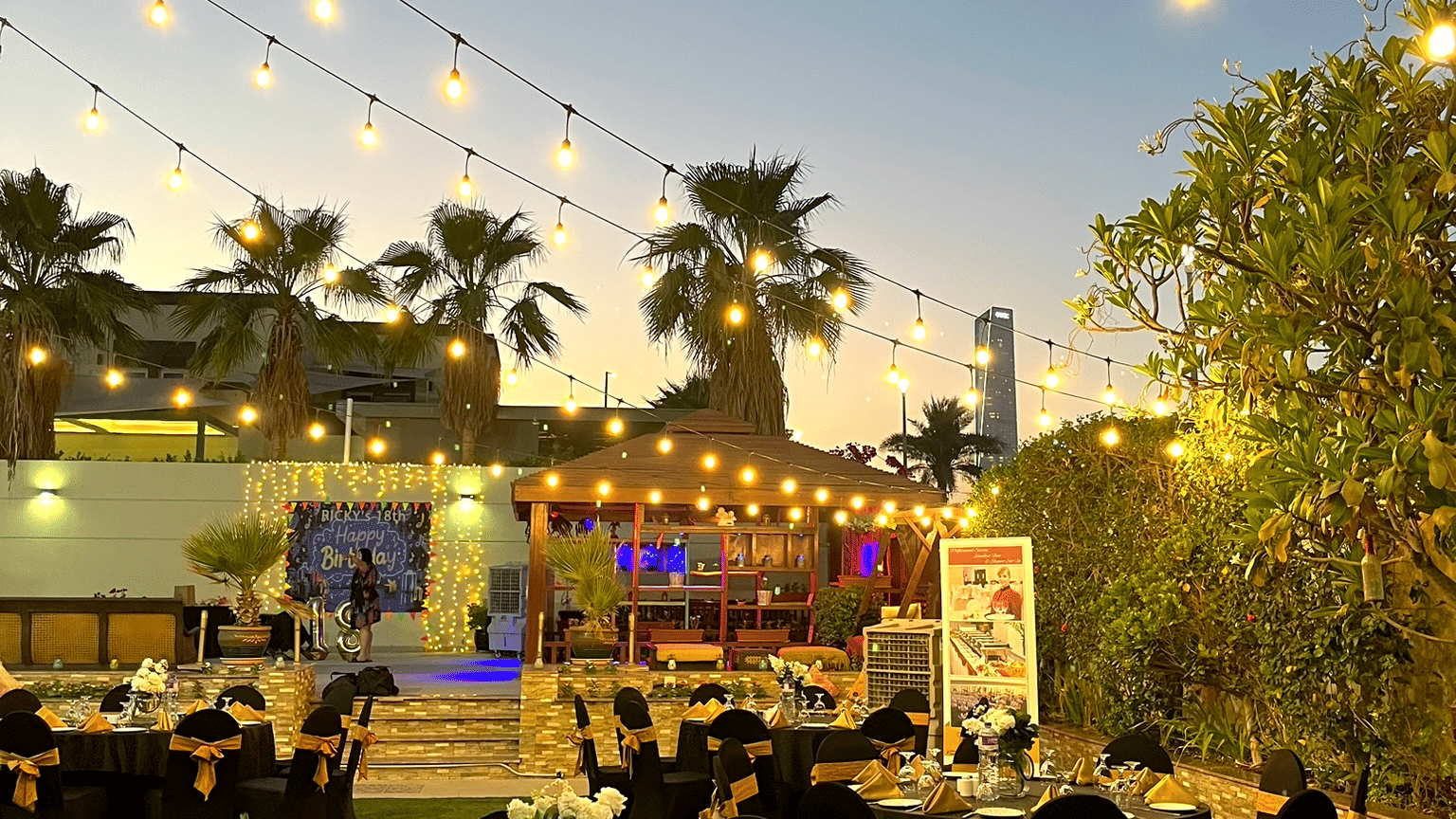 Outdoor event setup with round tables and chairs under string lights at Marco Polo Hotel.