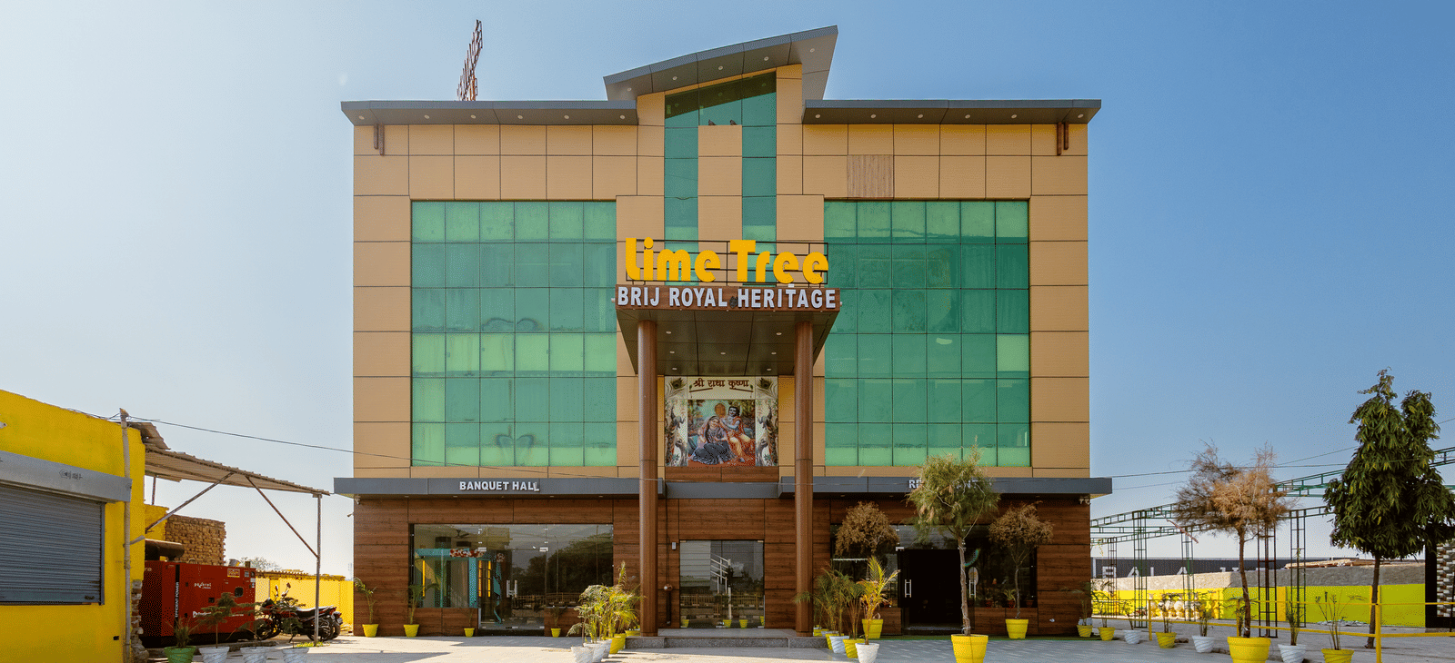 A daytime view of Lime Tree Hotels & Banquet, Vrindavan building with glass panels, yellow accents, and an open parking area in front.