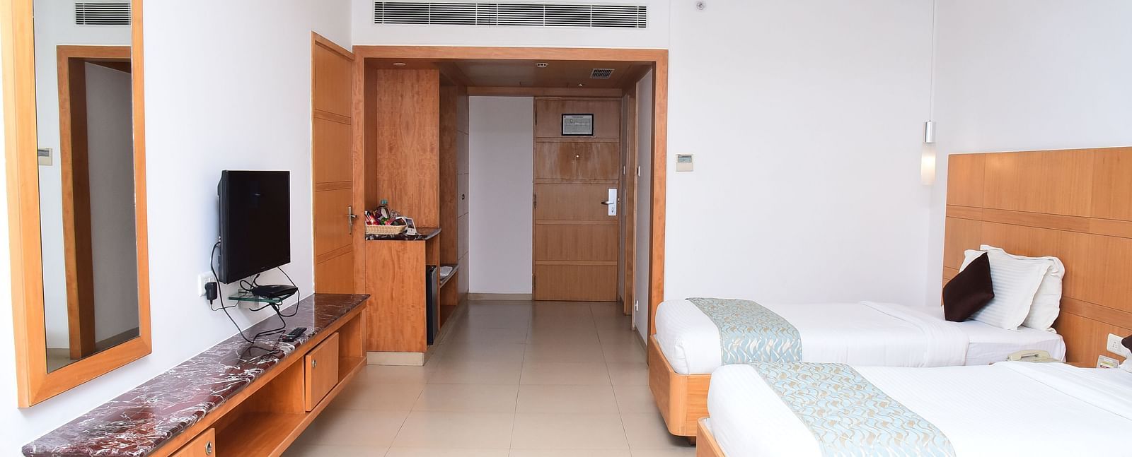 A well-furnished hotel room with twin beds, wooden interiors, and a sleek TV unit for added convenience.- Dhanalakshmi Srinivasan Hotels, Perambalur