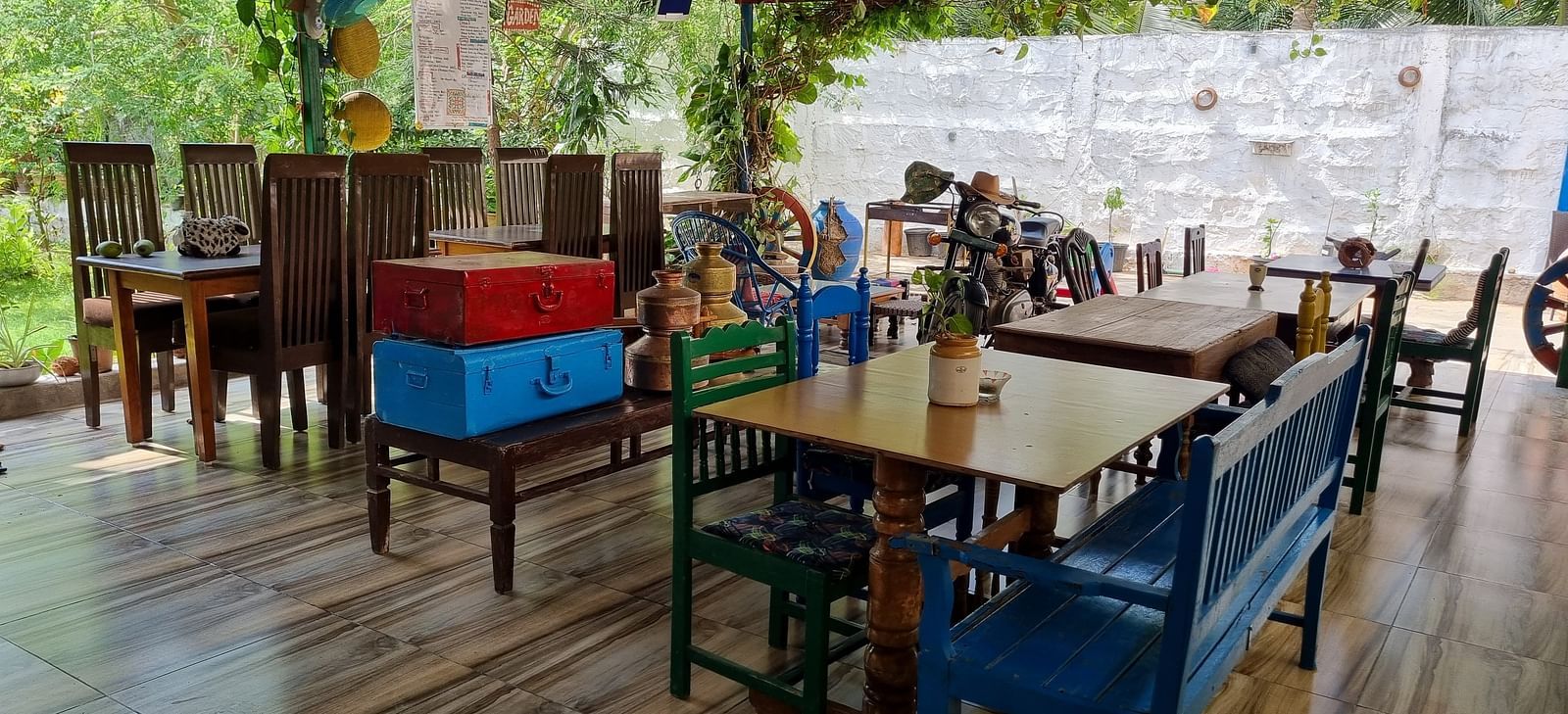 An open air dining area with wooden tables and chairs covered with a tin shed and a lush green garden in the background at Dream Vision Guest House.